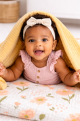 Load image into Gallery viewer, Baby girl wearing a pink ruffled outfit and white headband, peeking out from under a burpnbaby mango muslin blanket with a flower blanket underneath.